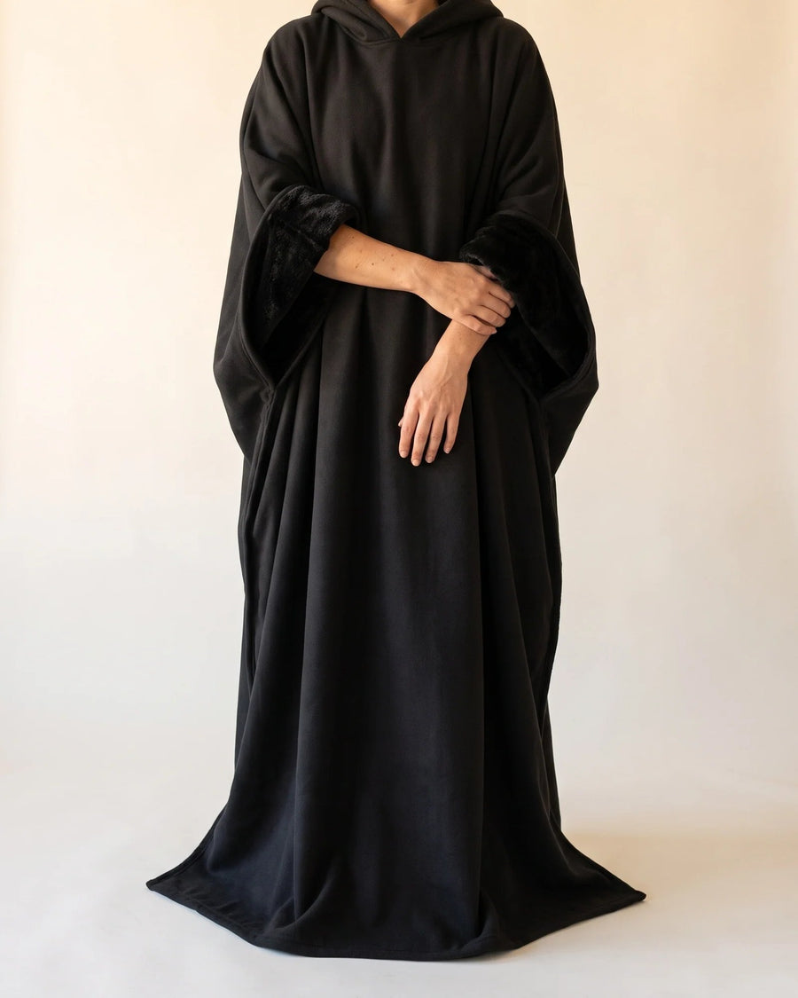 Woman in the black Hush blanket hoodie, a floor-length hooded cloak with plush faux fur at the sleeves