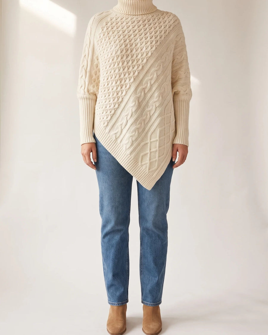 Cream Maple cable poncho sweater with a turtleneck and diagonal hem, worn with blue jeans